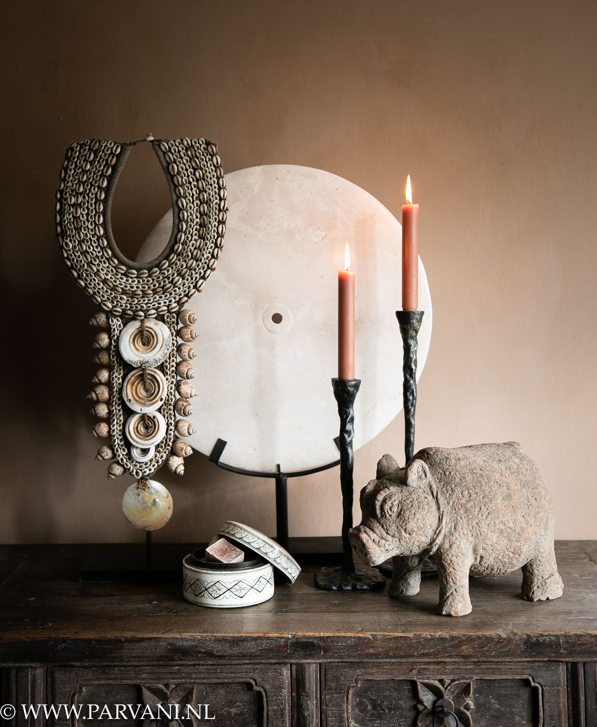 Grote ronde beige schijf op standaard met hoge schelpenketting met aardewerk varken, donkere bronzen kandelaren en zwart-wit ingelegd doosje