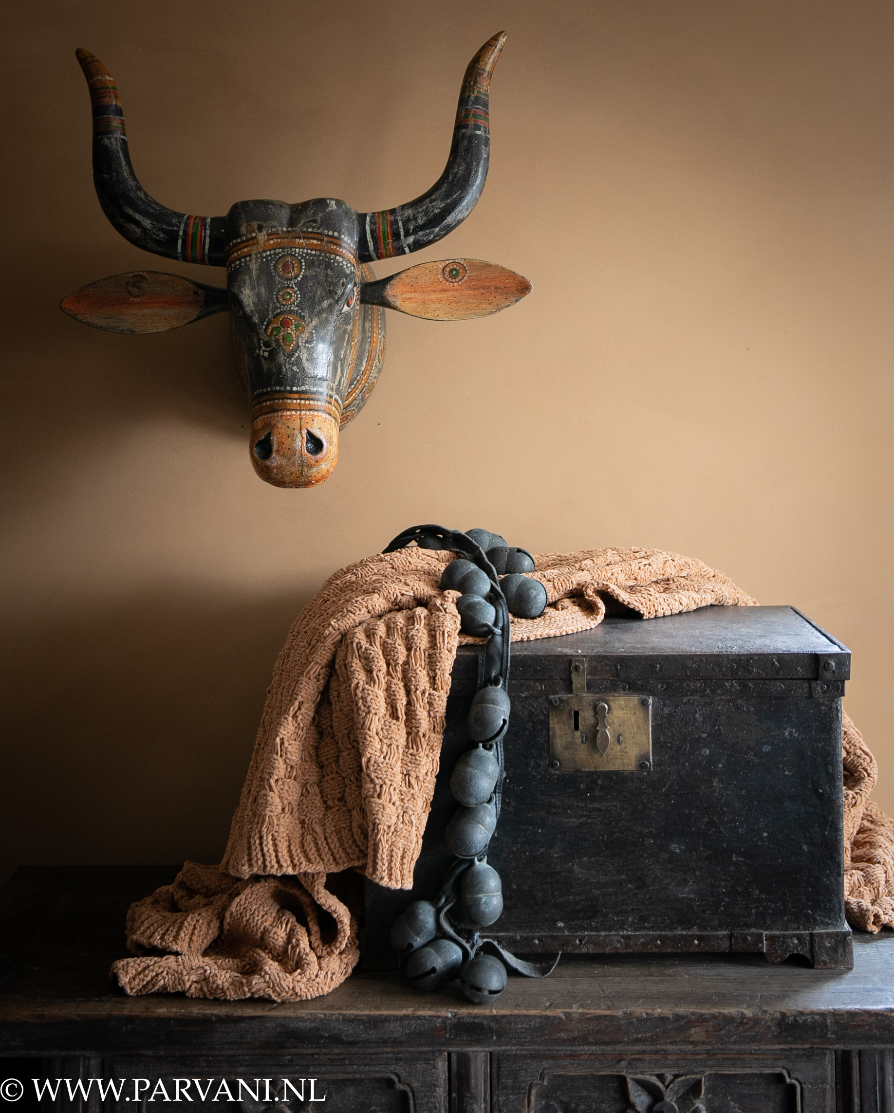 Oude zwarte metalen kist, riem met bronzen bellen en groot beschilderd koeienhoofd nandi uit India en camel kleurig gehaakt plaid