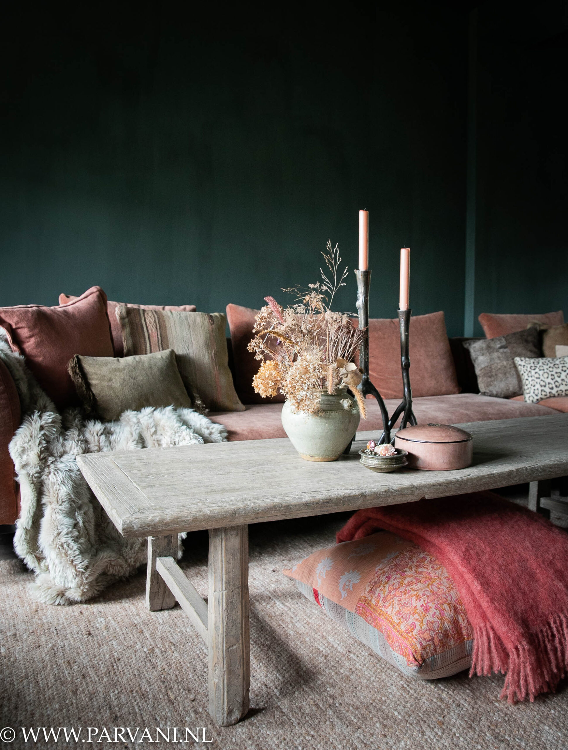 Lange smalle oude salontafel van licht naturel kleurig hout met houten poot, terra roze bank, vloerkussen en plaid met sierkussens