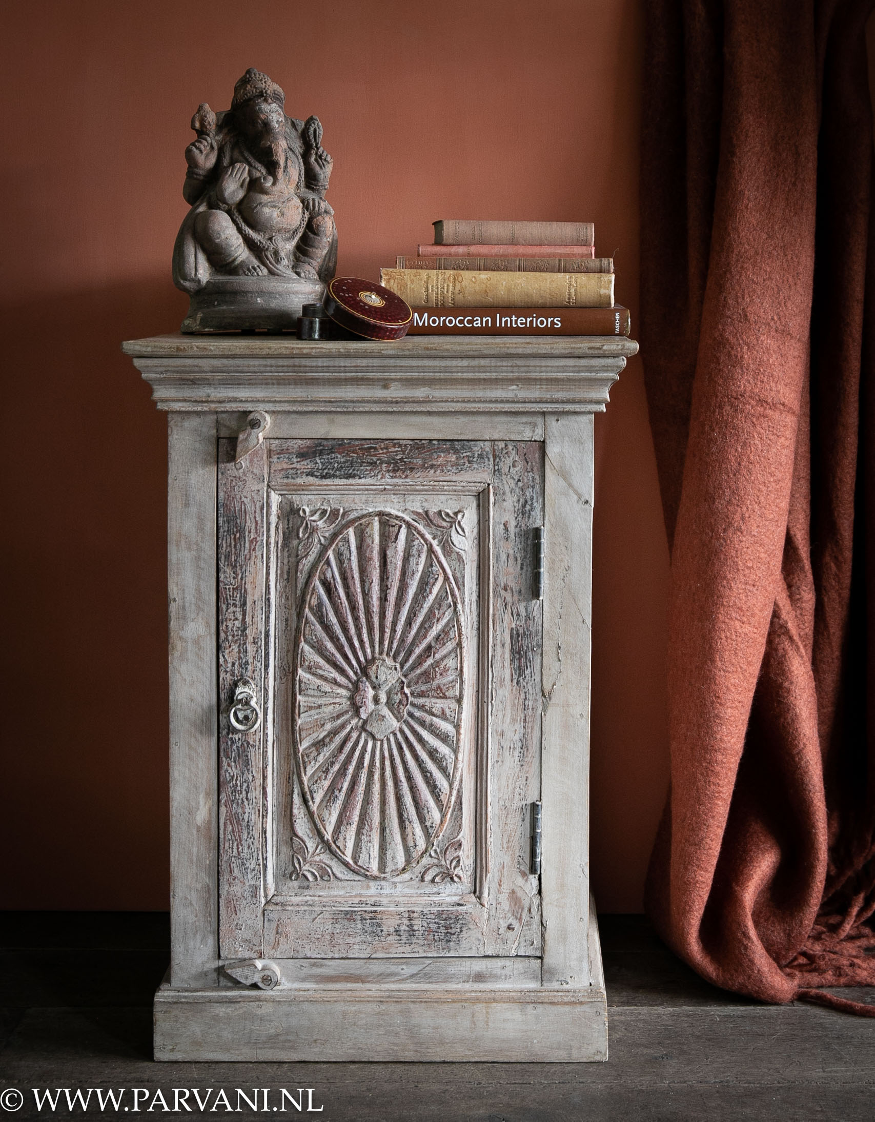 Oud houten kastje nachtkastje uit India met deurtje en snijwerk tegen warme terracotta muur met plaid en aardewerk Ganesha beeldje