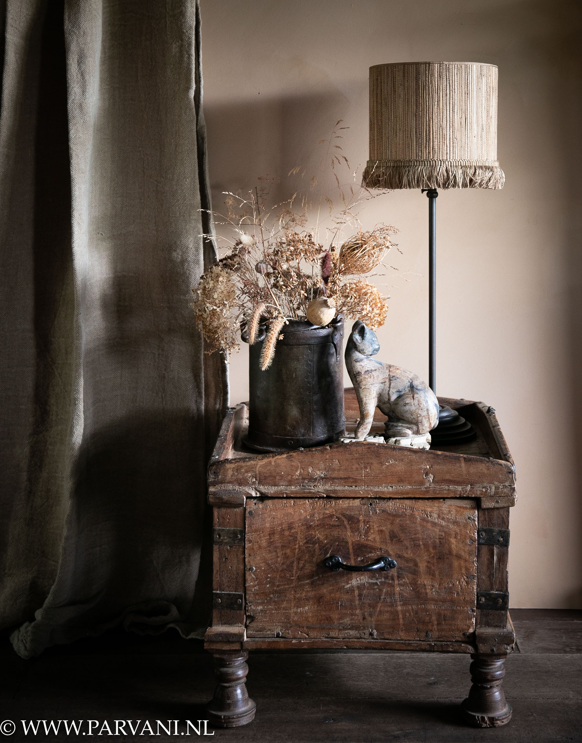 Vierkante houten bijzettafel met lade - nachtkastje uit India met tafellamp met raffia kapje en linnen Hoffz gordijn Zita en versteend houten kat met ijzeren vaas