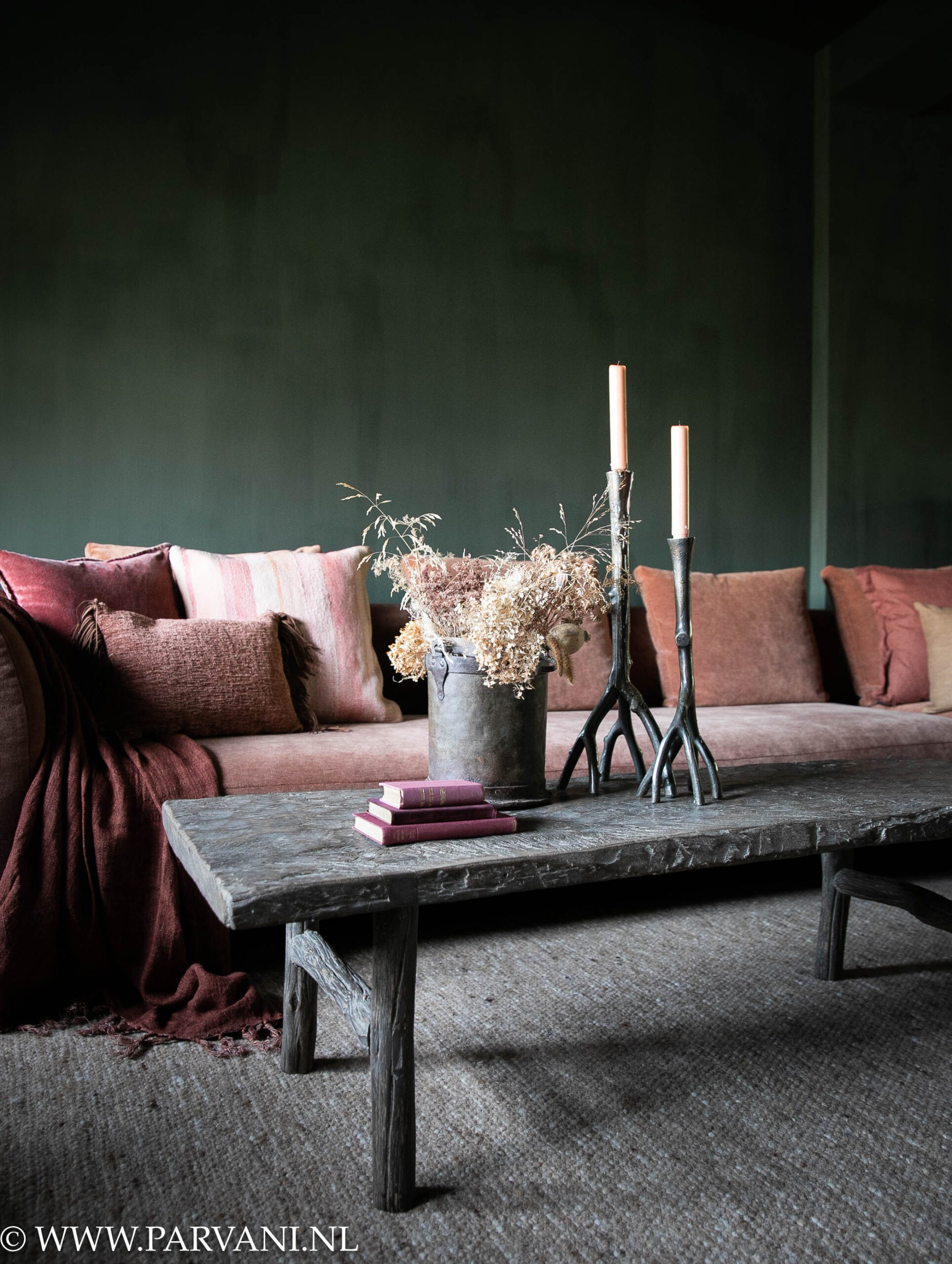 Smalle oude salontafel in donkere bruingrijze kleur met doorleefd blad en houten onderstel bij oranje bank met roze kussens